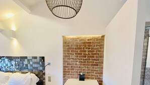 A modern bedroom with a brick accent wall, a hanging light fixture, a bedside table, and a bed with white bedding. - Hostellerie la Fontaine (Saint-Paul-de-Vence)