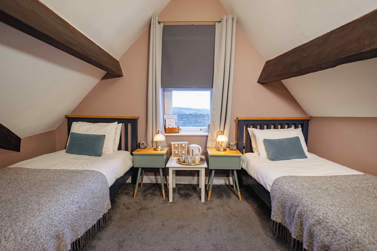 A cozy attic bedroom with two beds, a small table with lamps, and a window with a view.