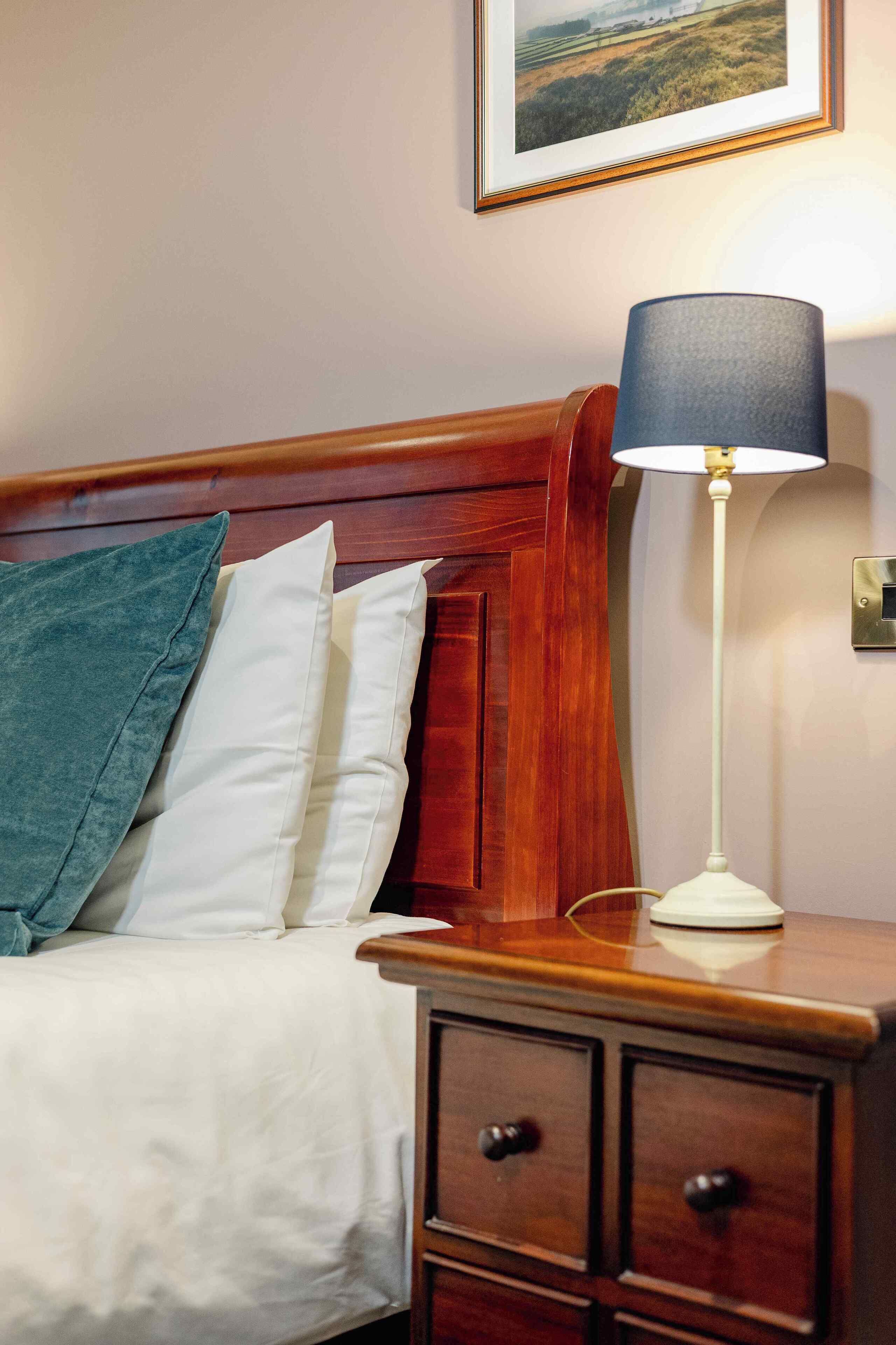 A neatly made bed with a white and teal pillow, a wooden nightstand with a lamp, and a framed picture on the wall.