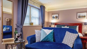 Una habitación con un sofá azul, una pequeña mesa redonda con un libro y una copa de vino, un espejo, una ventana con cortinas, una cama con cabecero de madera y dos lámparas de pared.