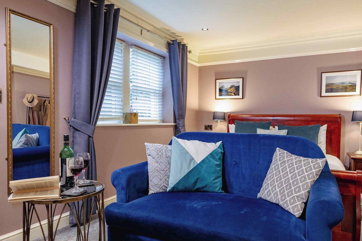 A room with a blue sofa, a small round table with a book and a glass of wine, a mirror, a window with curtains, a bed with a wooden headboard, and two wall-mounted lamps.