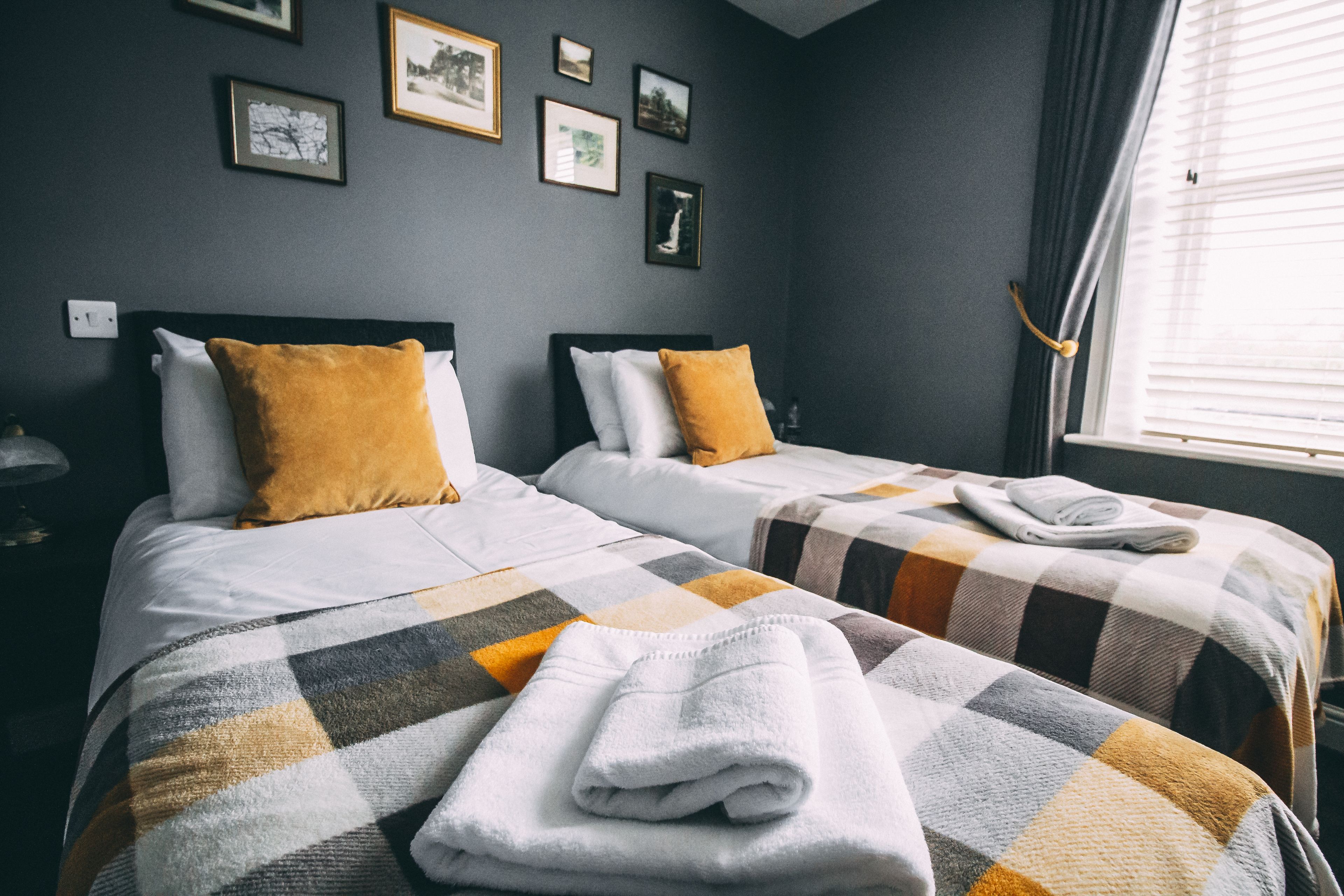 A hotel room with two beds, each with a checkered blanket and a folded towel on the bed. There are framed pictures on the wall and a window with curtains.