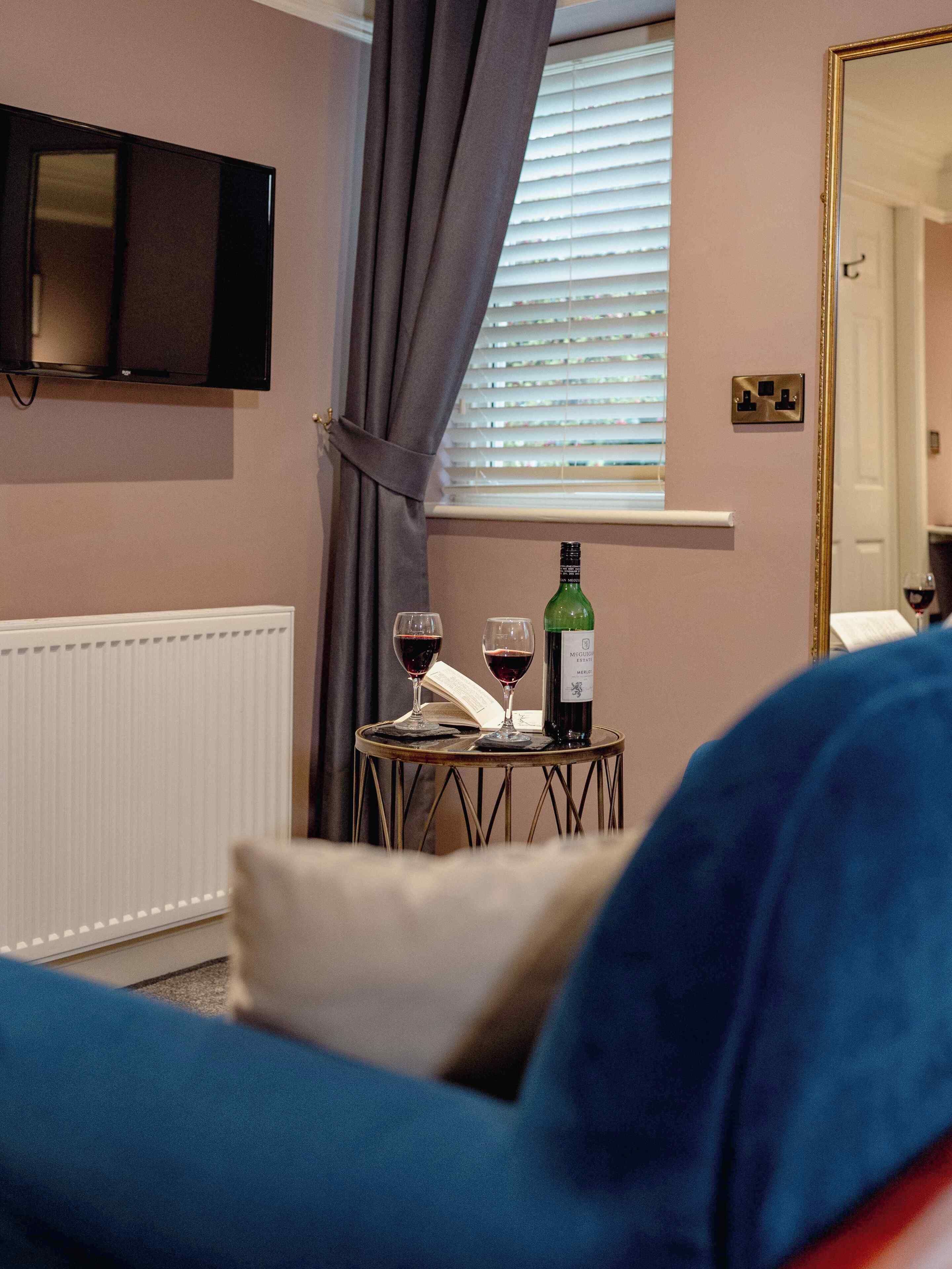 A room with a television mounted on the wall, a window with blinds, a table with two wine glasses and a bottle, and a blue couch.