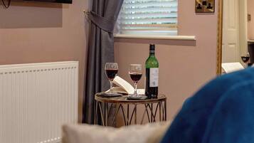 A room with a television mounted on the wall, a window with blinds, a table with two wine glasses and a bottle, and a blue couch.