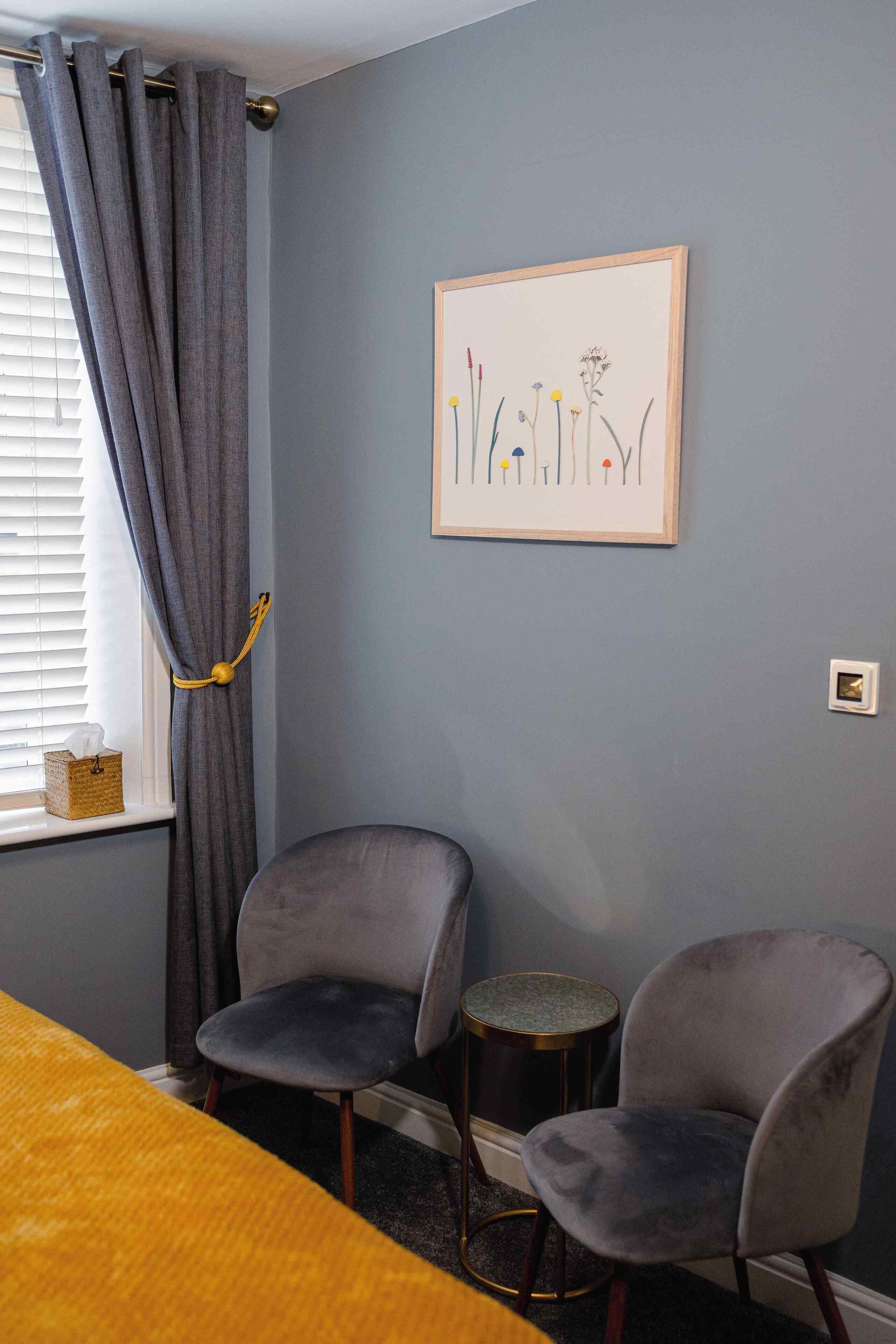 A room with two grey armchairs, a small round table, a yellow bed, and a window with grey curtains.