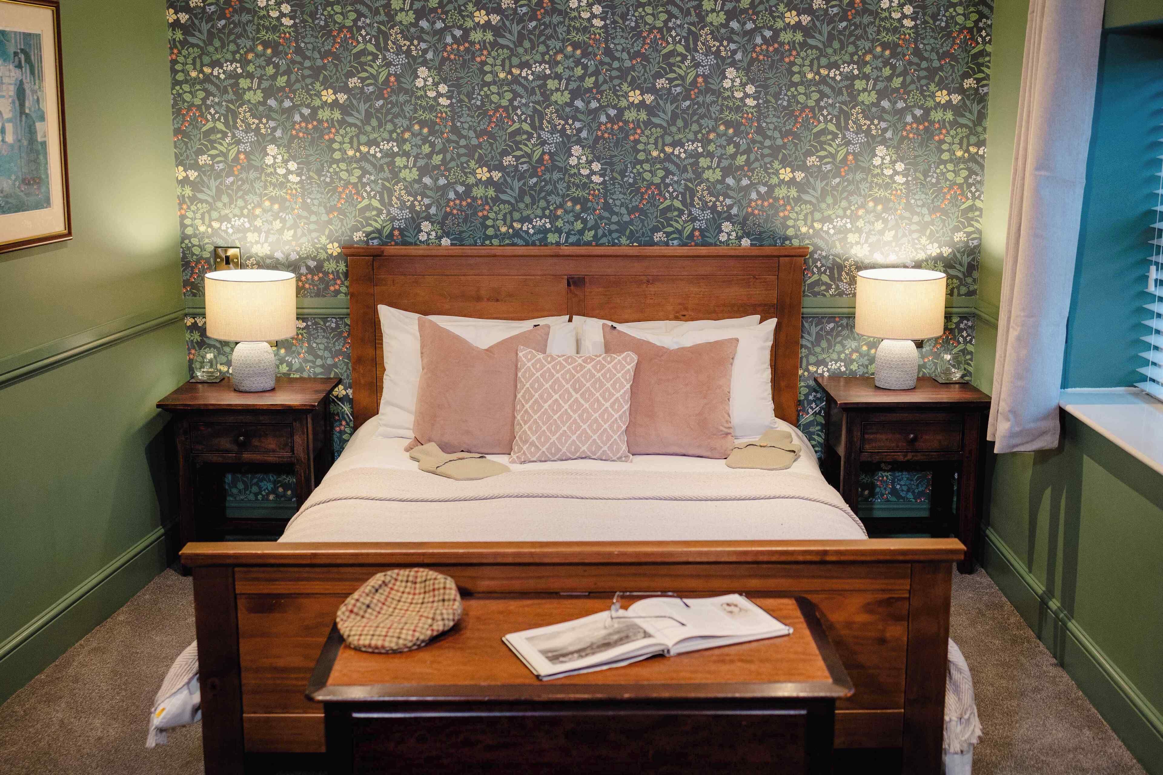 A neatly made bed with a wooden headboard, two bedside lamps, and a small table with a book and a hat.