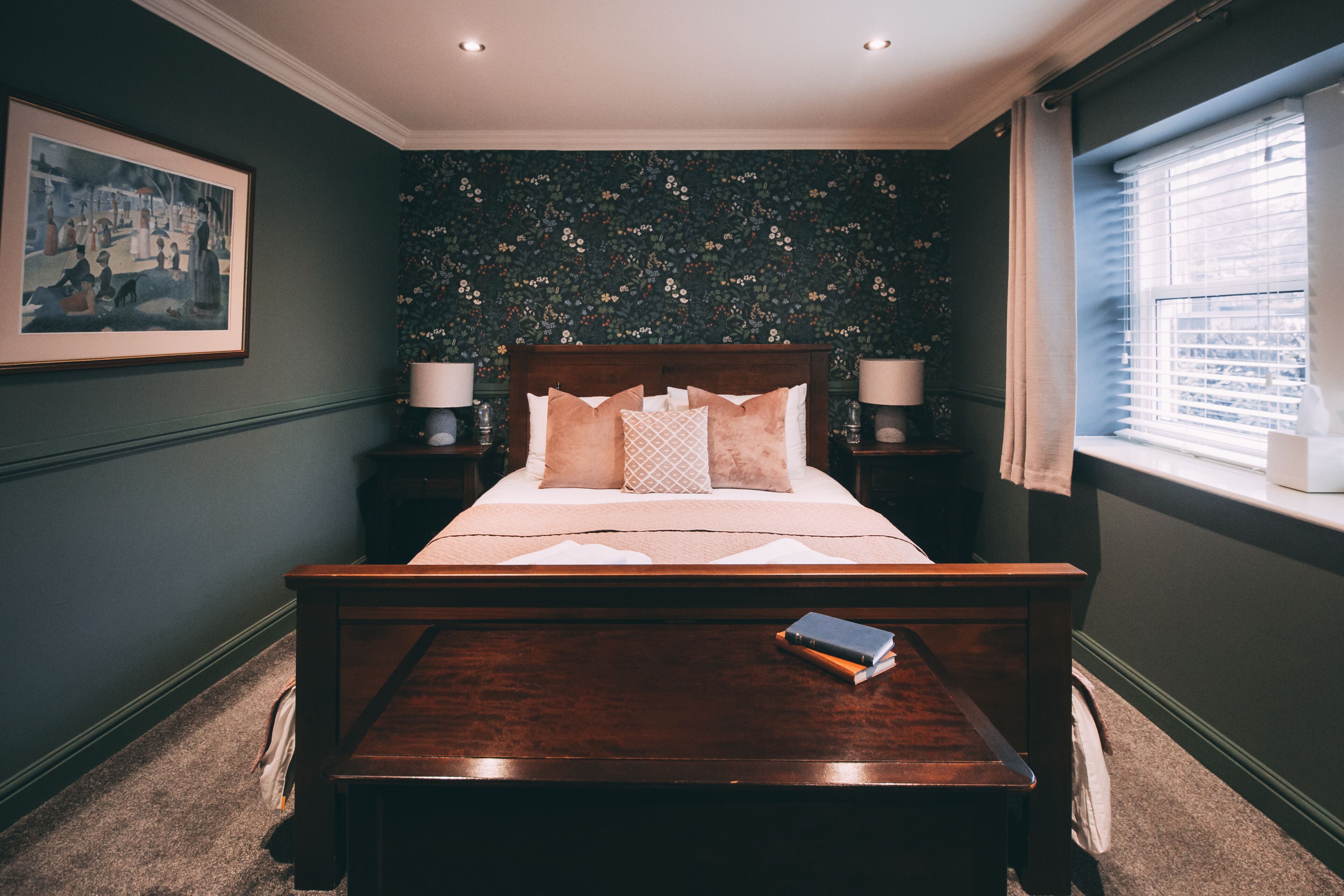A bedroom with a large bed, two bedside tables with lamps, a framed picture on the wall, and a window with blinds.