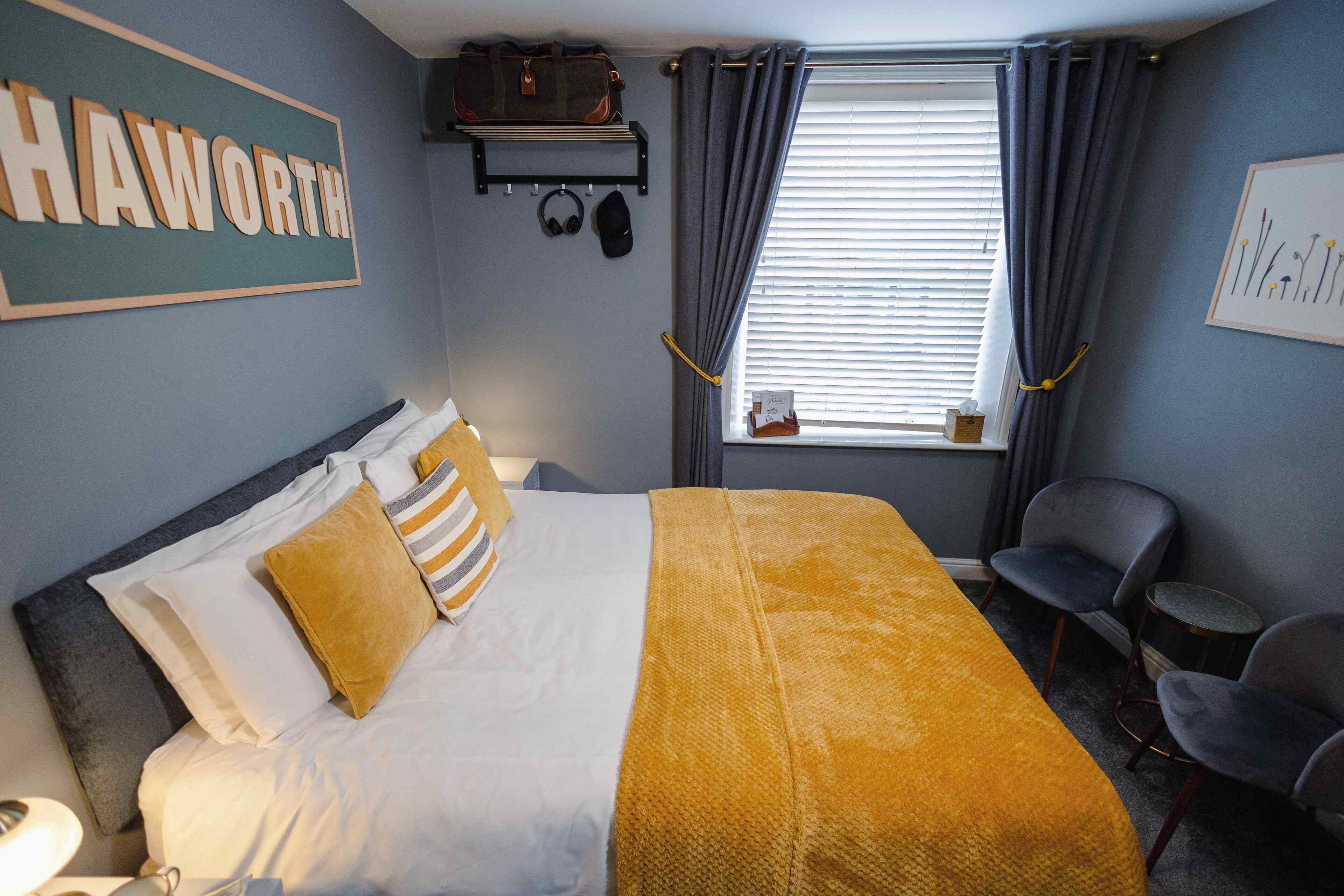 A hotel room with a bed, a headboard, a bedside table, a chair, a window with curtains, a wall-mounted shelf with a bag and headphones, and a framed picture on the wall.