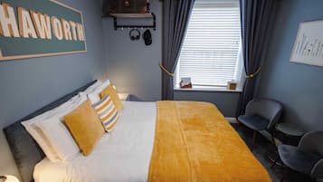 A hotel room with a bed, a headboard, a bedside table, a chair, a window with curtains, a wall-mounted shelf with a bag and headphones, and a framed picture on the wall.