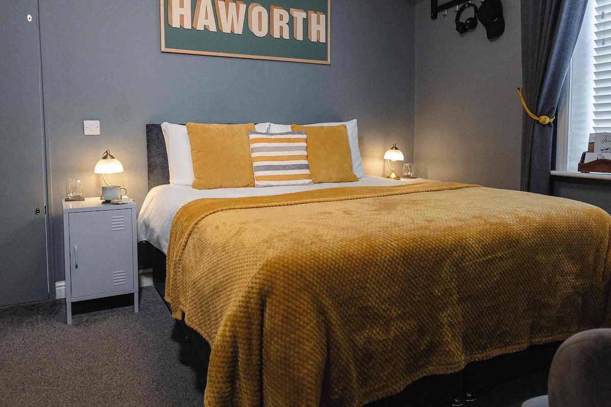 A bedroom with a bed, nightstands, a wall-mounted shelf with hats, a sign reading "HAWORTH", and a window with curtains.