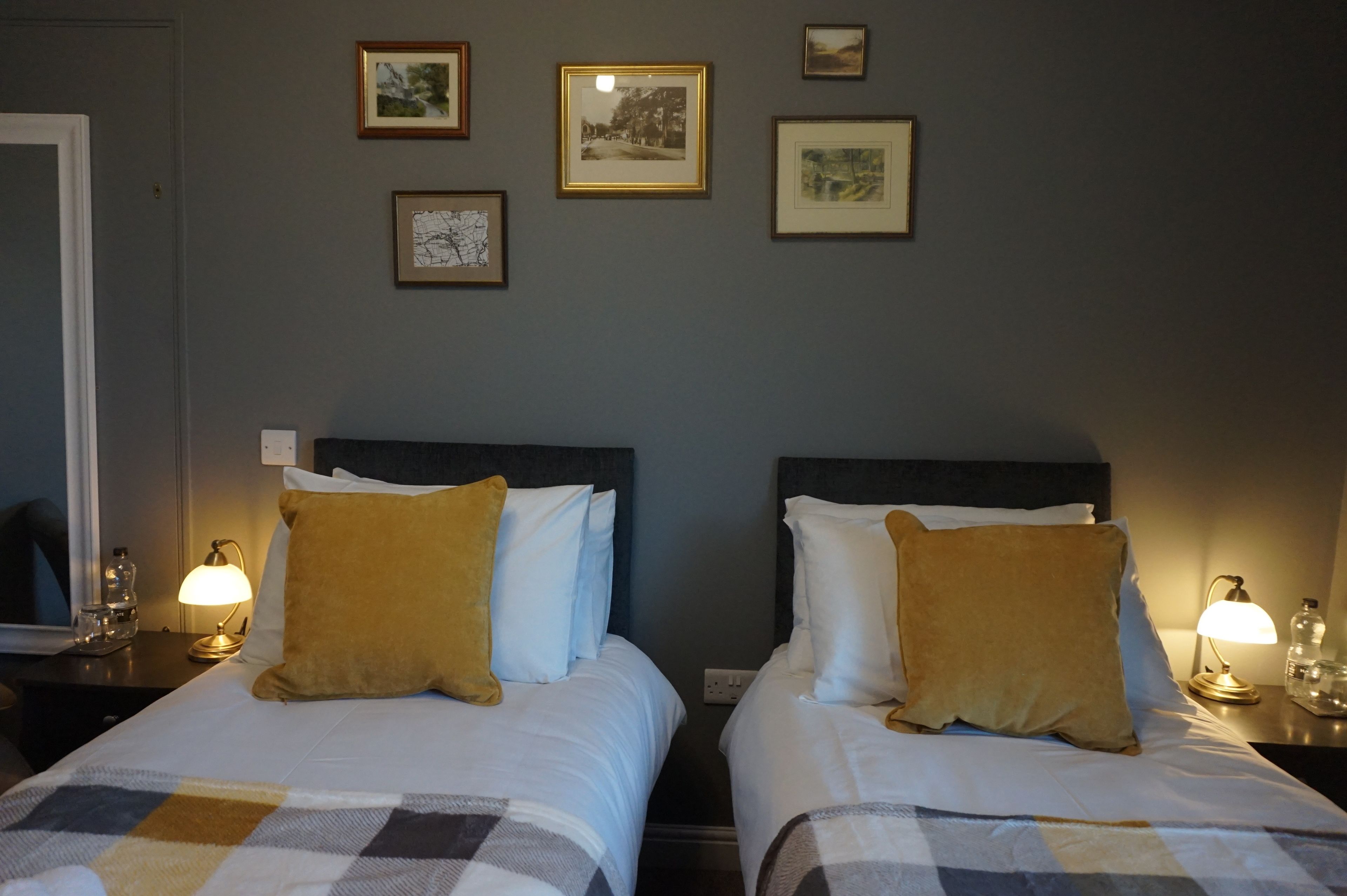 Habitación de hotel con dos camas, cada una con cabecera, mesita de noche con lámpara y cuadros en la pared.