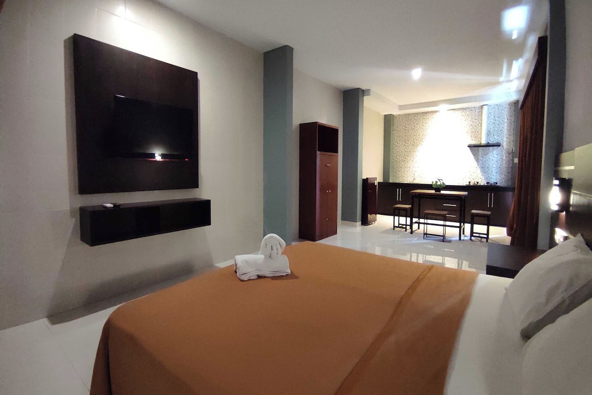 A hotel room with a bed, a flat-screen TV mounted on the wall, a small table with a plant, and a seating area with chairs.