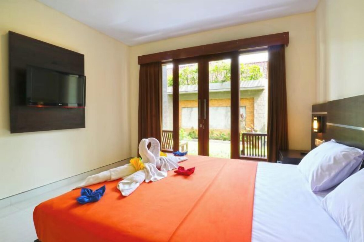 A hotel room with a bed, a TV mounted on the wall, and a balcony with a view of greenery.