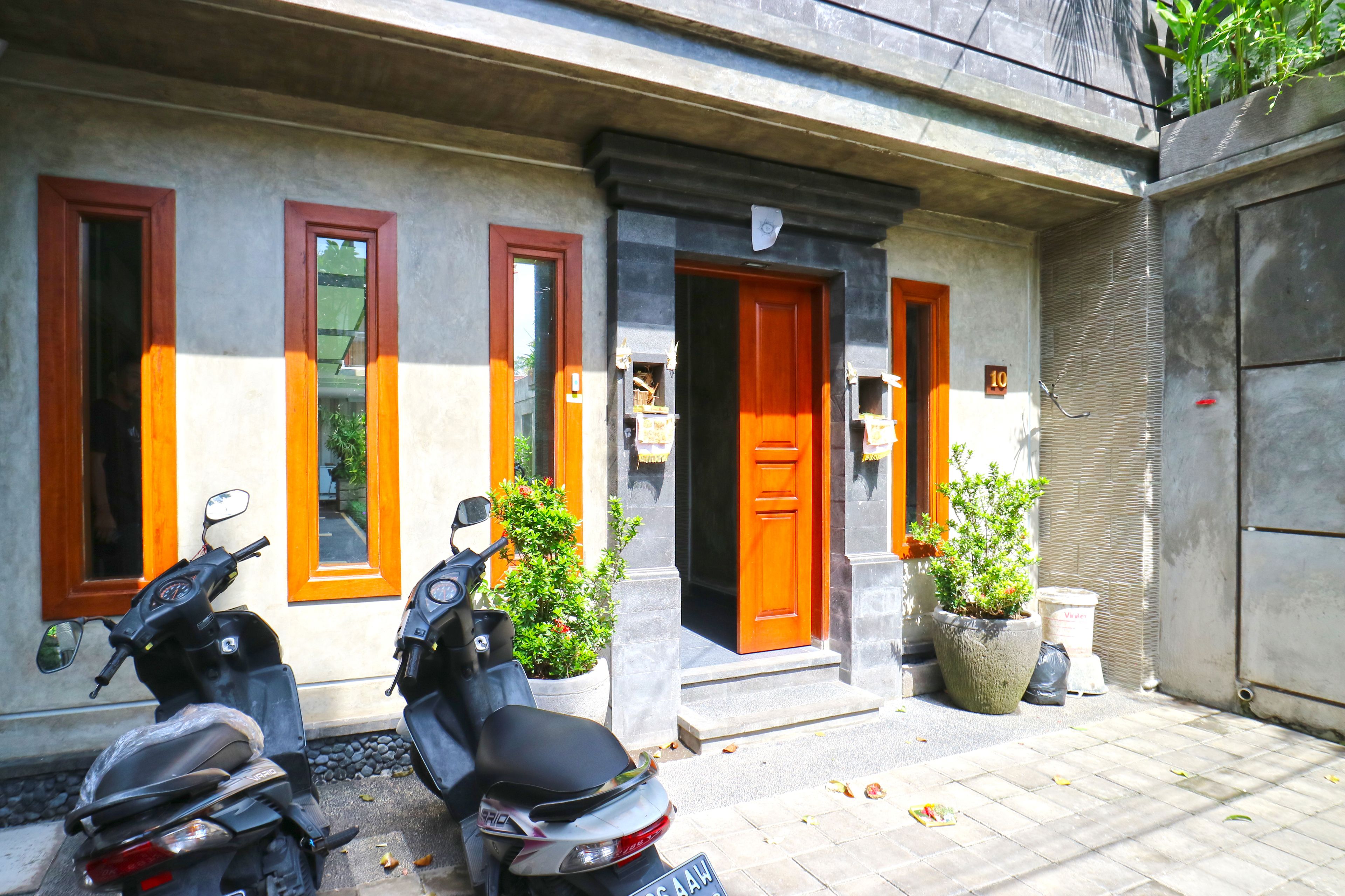 A building with a concrete facade, two wooden-framed windows, and a red door. There are two scooters parked in front, and potted plants are placed near the entrance.