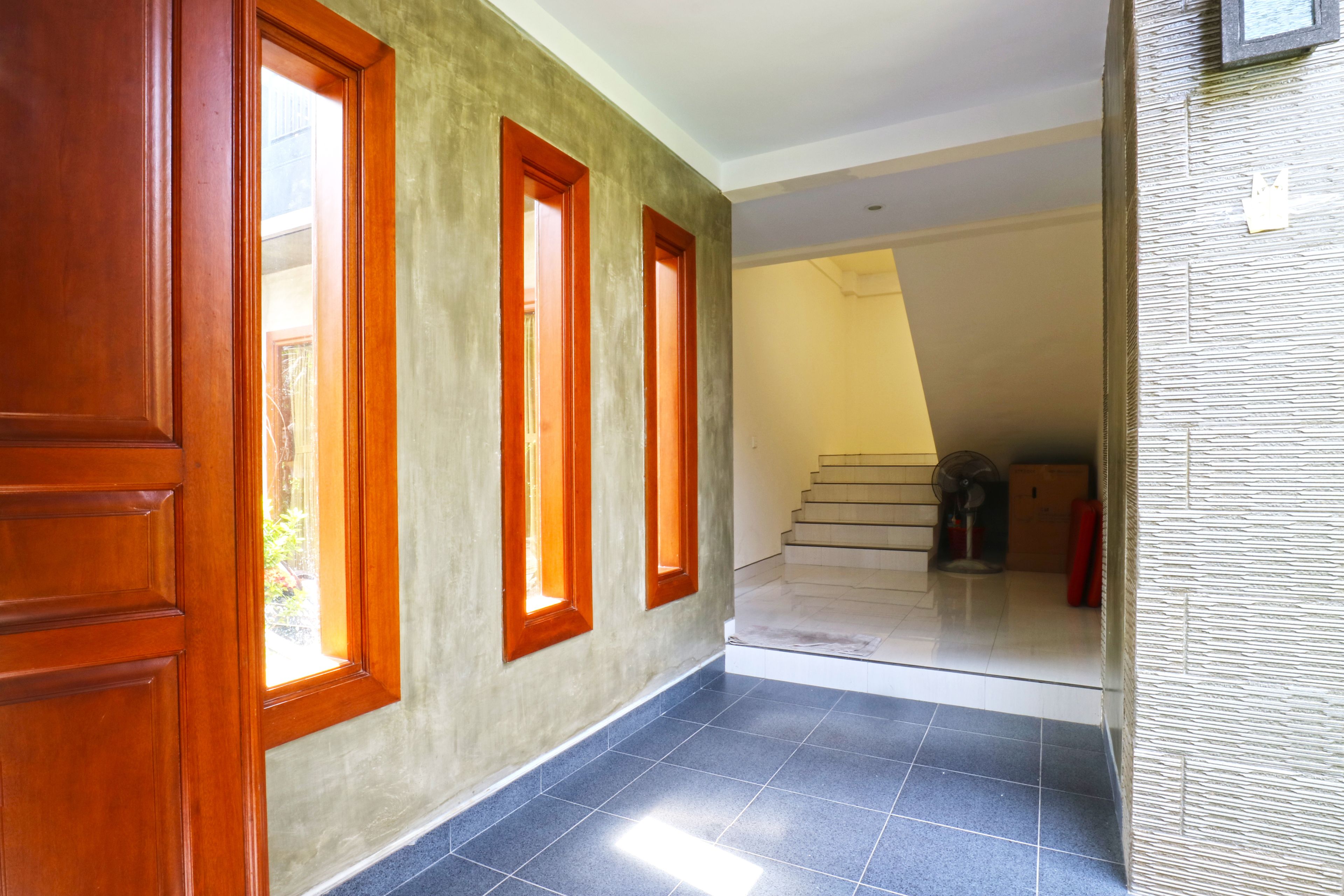 Un corridoio con una porta in legno, tre finestre con cornici rosse, un pavimento piastrellato e una scala che conduce a un piano superiore.