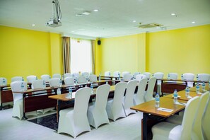 Une salle de conférence avec des rangées de chaises blanches, des tables et des bouteilles d’eau.
