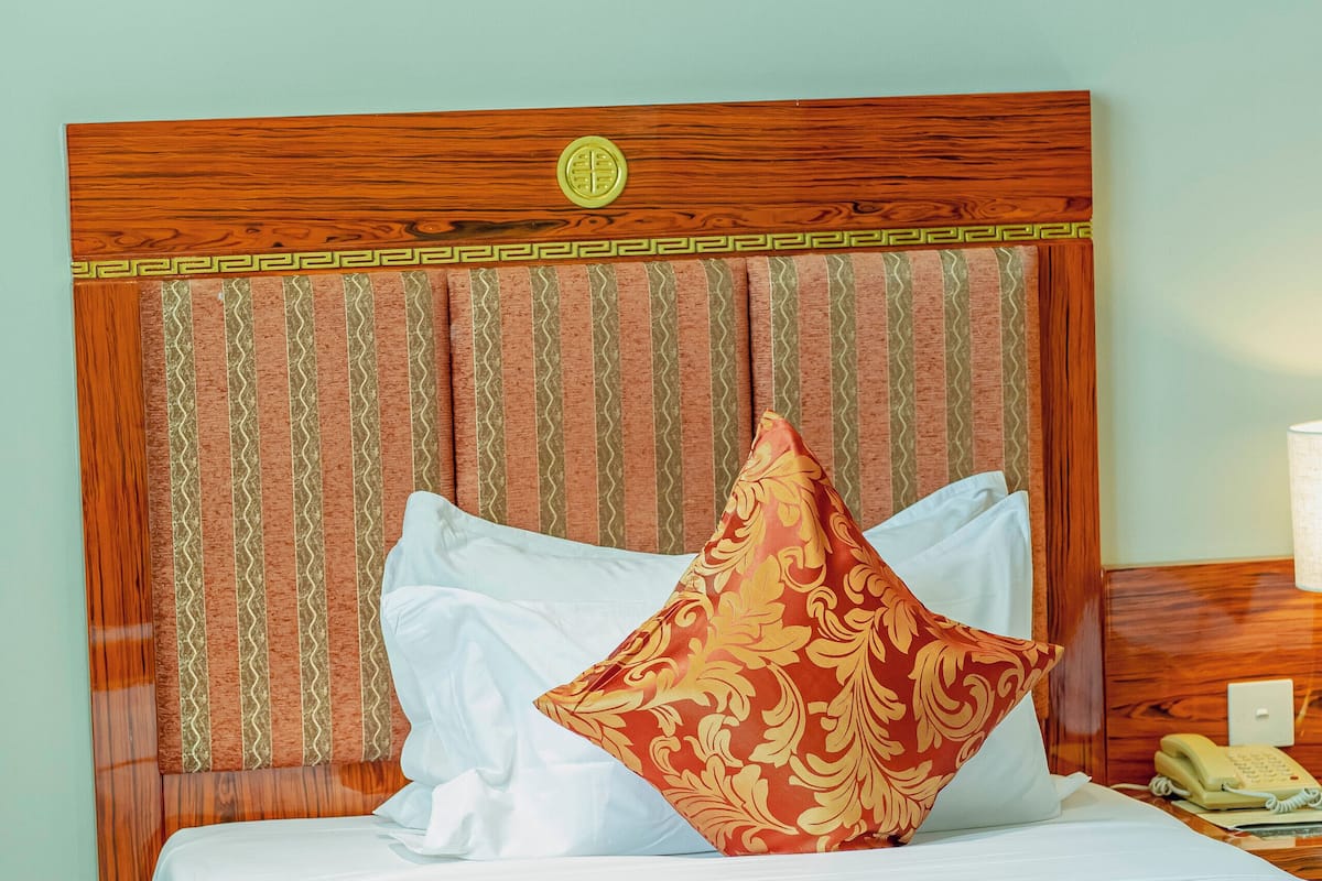Un lit bien fait, avec une tête de lit en bois, une literie blanche et un coussin rouge décoratif. On aperçoit une table de chevet avec un téléphone et un récipient en verre contenant des fruits.