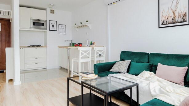 Una sala de estar moderna con un sofá verde, una mesa de centro de vidrio y una cocina con armarios blancos y un microondas.