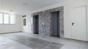 Habitación amplia con piso de baldosas, pared decorativa de mármol y dos puertas, una de ellas acceso al ascensor.