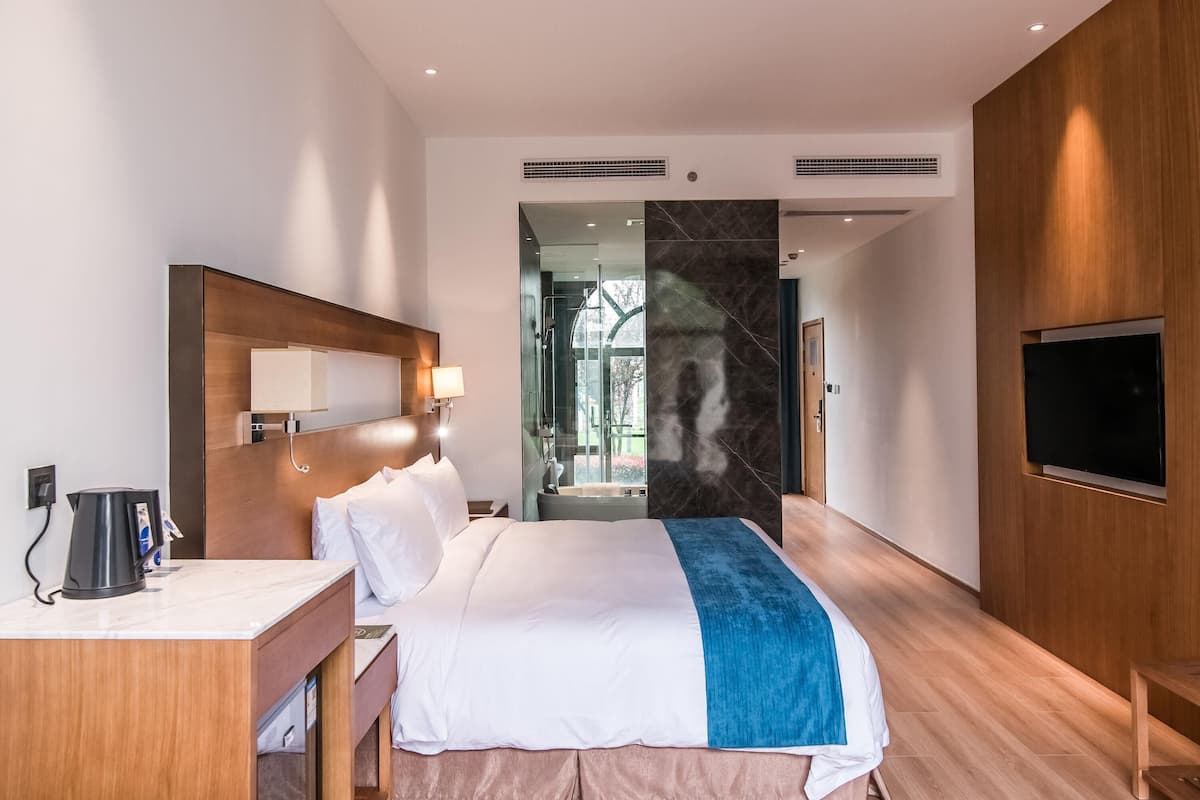 A modern hotel room with a bed, a desk, a coffee maker, a TV, and a bathroom visible through a glass door.