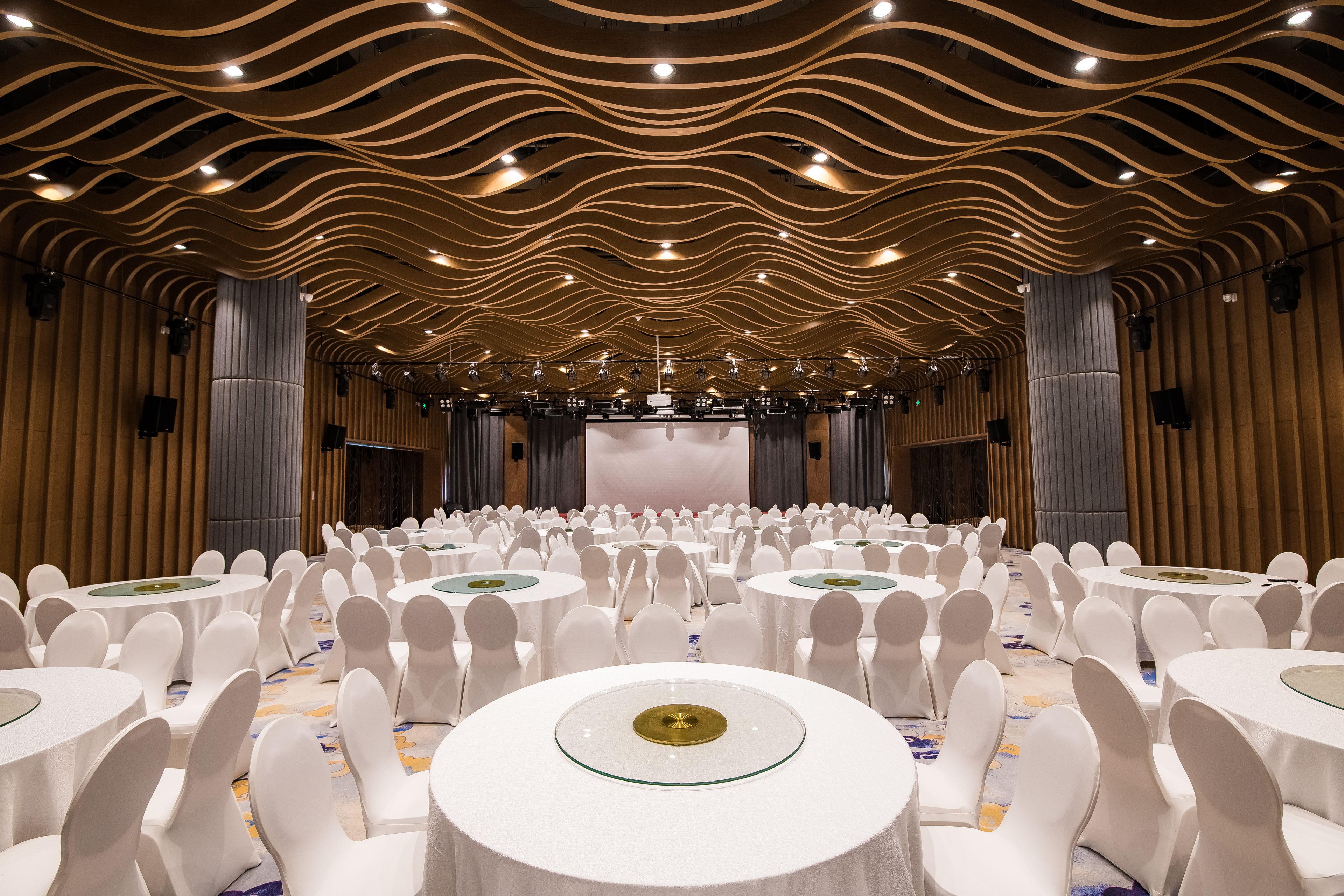 A banquet hall with round tables and white chairs, a stage at the front, and a wavy ceiling design.