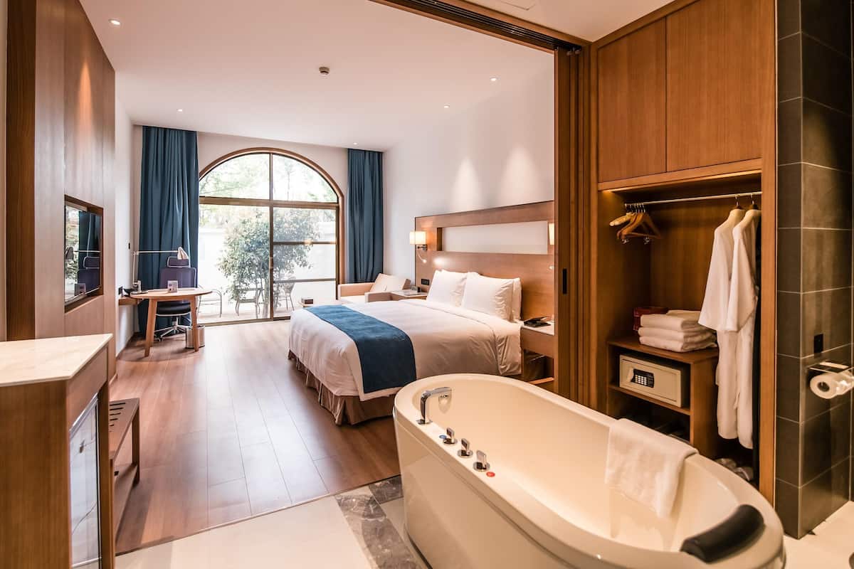 A modern hotel room with a large bed, a sitting area, a desk, a bathtub, and a wardrobe.
