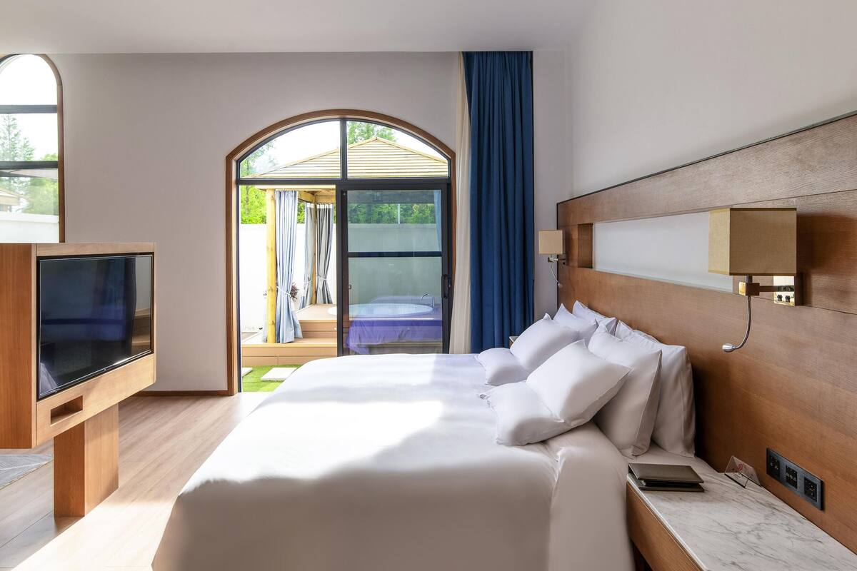 A hotel room with a bed, a TV, a desk, and a view of an outdoor area.