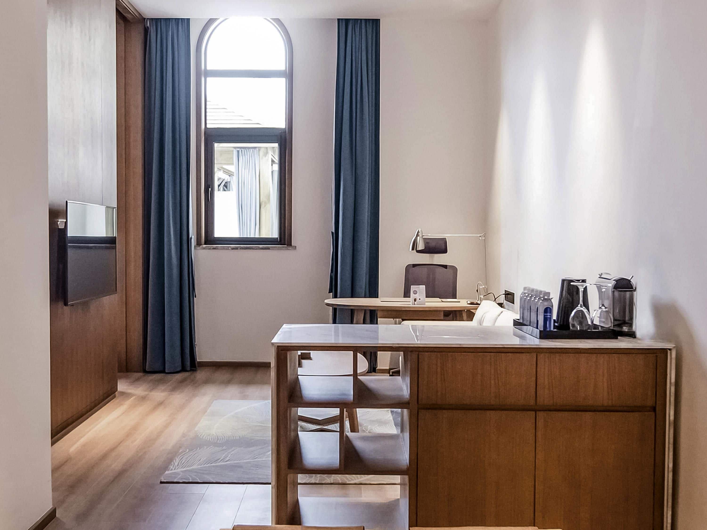 A compact hotel room with a wooden desk, a chair, a flat-screen TV mounted on the wall, a window with curtains, and a small kitchenette area.