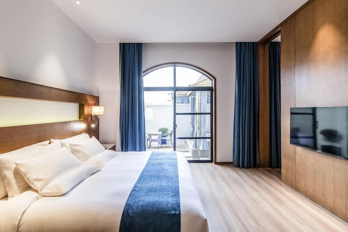 A hotel room with a large bed, a TV, a wooden headboard, and a view through a large window.