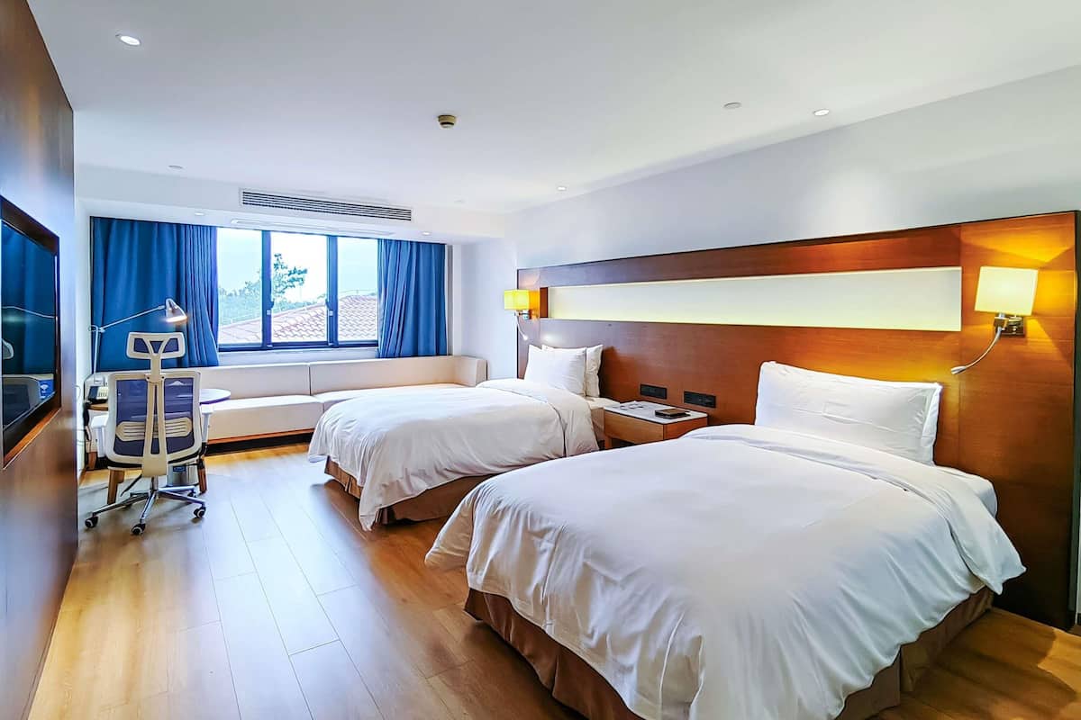 A hotel room with two beds, a desk, a chair, a TV, and a window with curtains.
