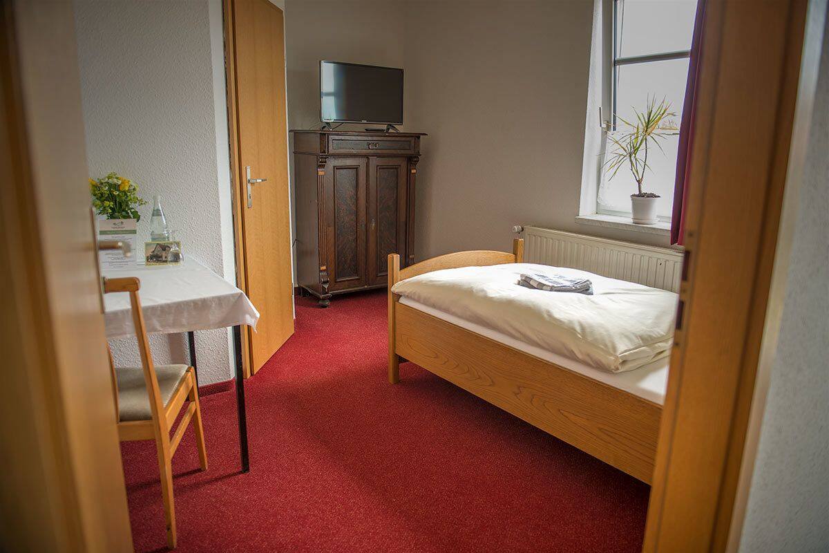 Une petite chambre avec un lit simple, une armoire en bois, un téléviseur posé sur un meuble, une petite table avec une chaise et une fenêtre agrémentée d’une plante.