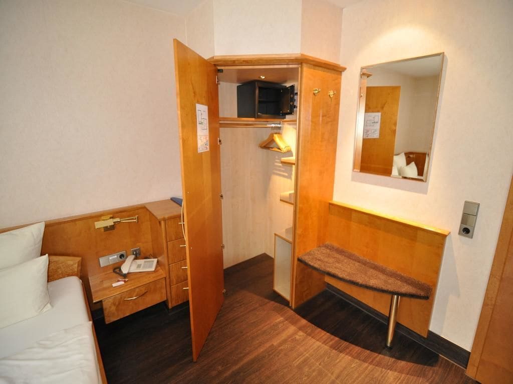 A hotel room with a bed, a wooden desk, a bench, a wardrobe, a mirror, and a small shelf.