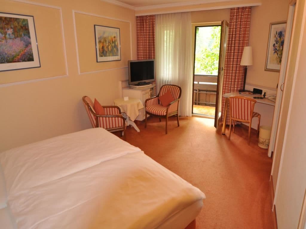 A hotel room with a bed, a desk with a television, two striped chairs, a small table, and a window with curtains.