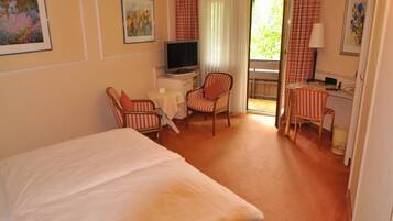 A hotel room with a bed, a desk with a television, two striped chairs, a small table, and a window with curtains.