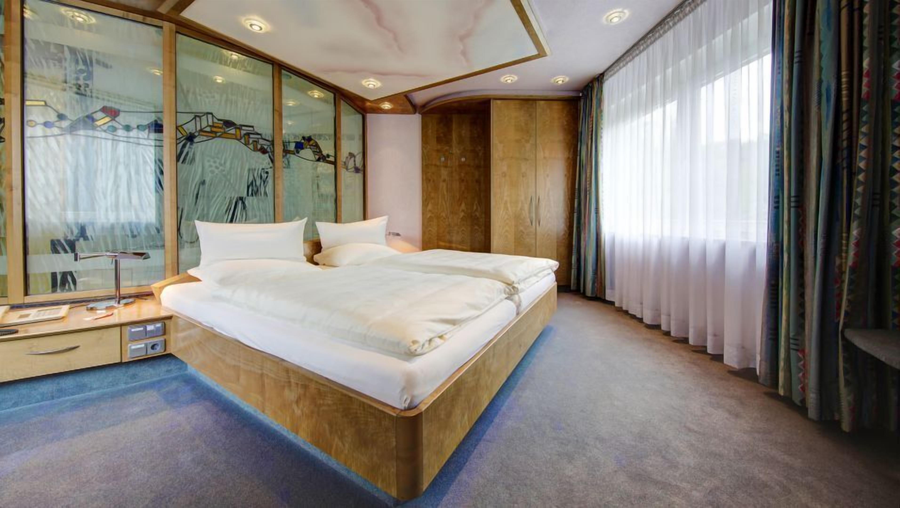 A modern bedroom with a large bed, a wooden headboard, a glass wall with artwork, and a large window with curtains.