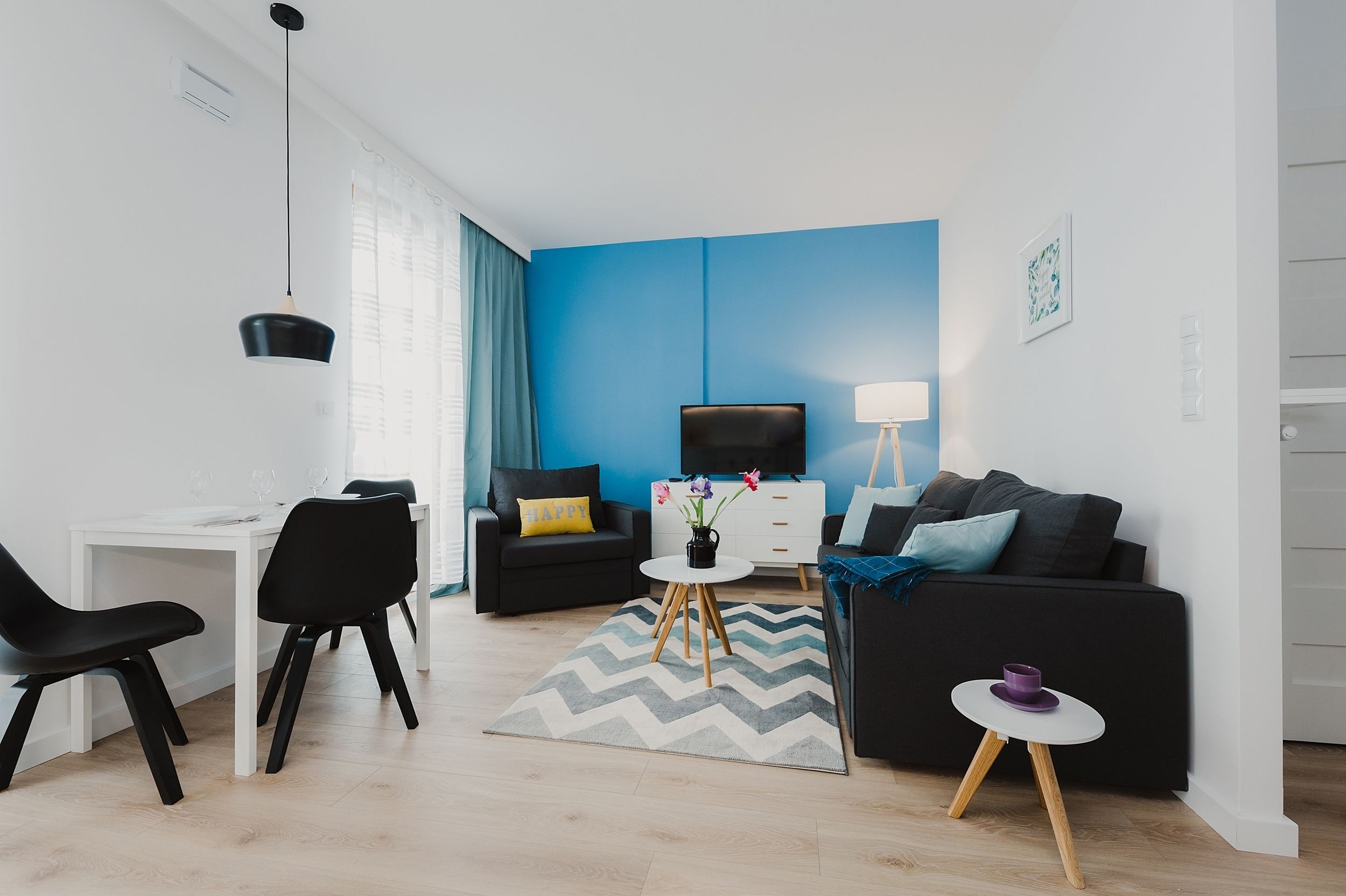 Un salon moderne avec un mur d’accent bleu, un canapé noir, un bureau blanc et une petite table ronde sur laquelle se trouve une tasse violette.