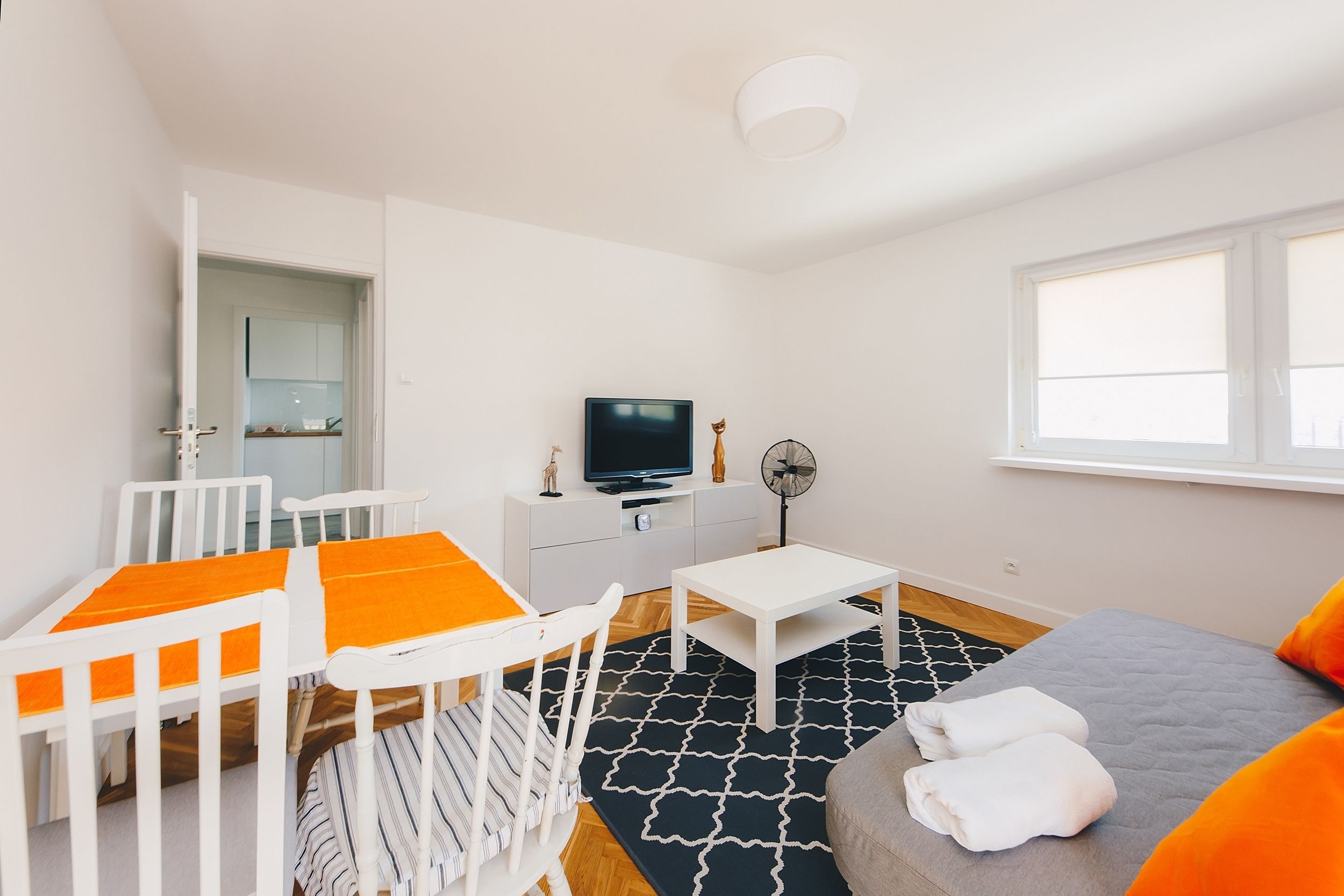 Une pièce moderne et bien éclairée, avec un lit blanc, un canapé gris, une petite table blanche, un téléviseur posé sur un meuble blanc et un tapis aux motifs noir et blanc.