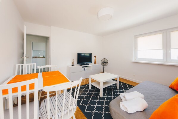 Une pièce moderne et bien éclairée, avec un lit blanc, un canapé gris, une petite table blanche, un téléviseur posé sur un meuble blanc et un tapis aux motifs noir et blanc.