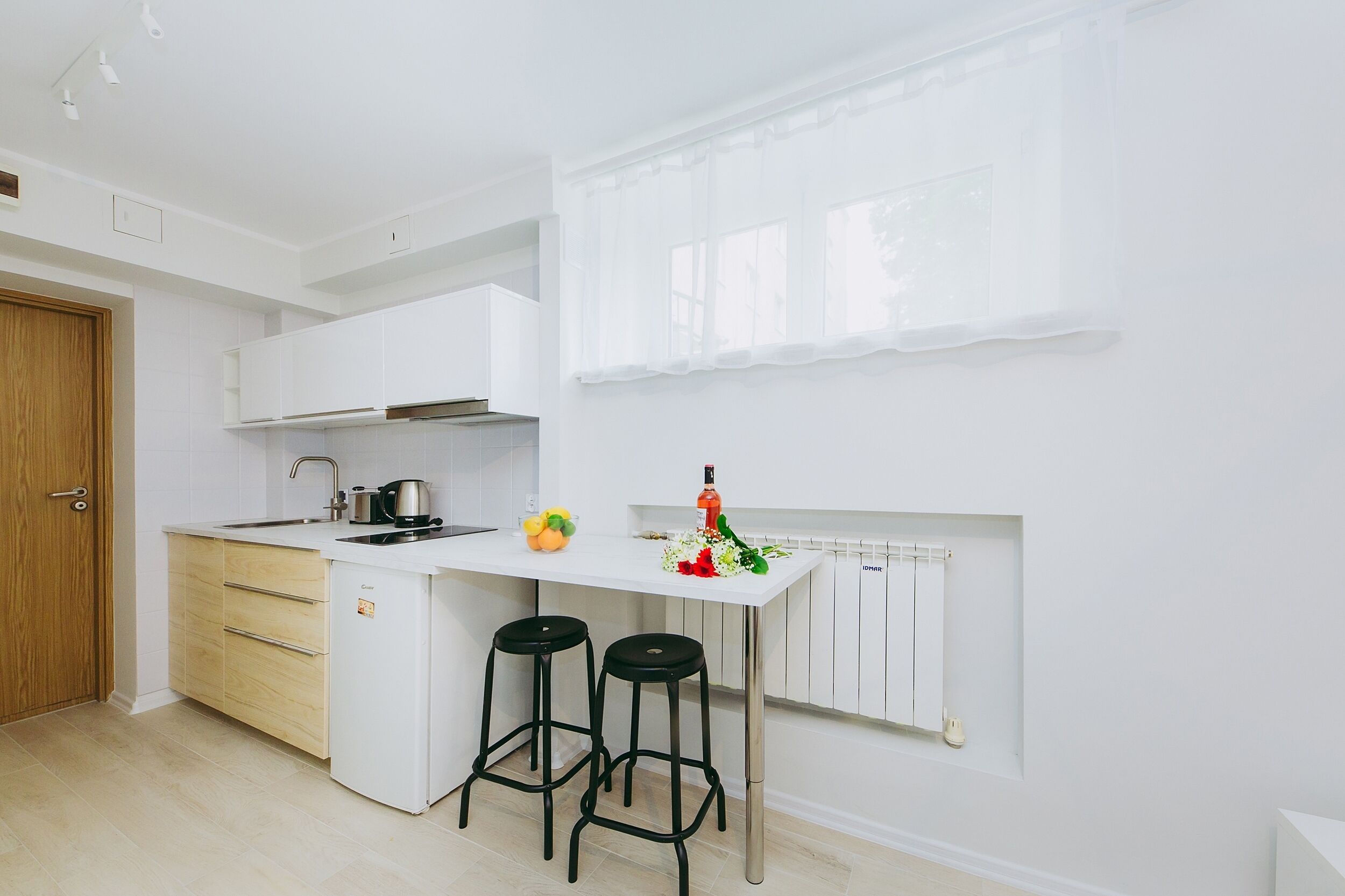 A modern kitchen with white cabinetry, a white countertop, two black stools, and a window with sheer curtains.