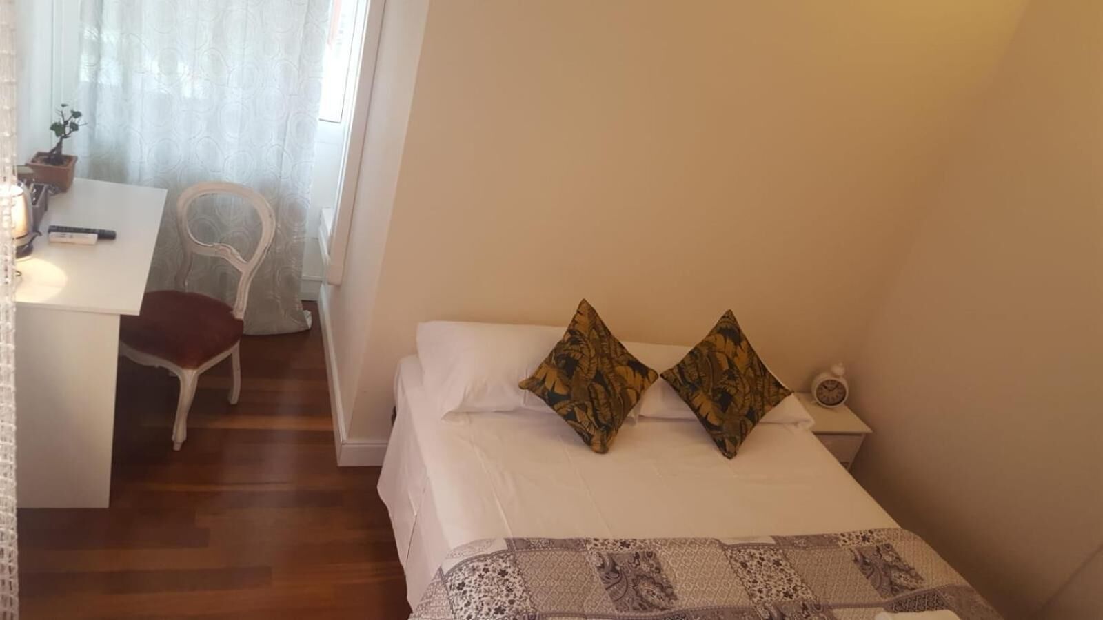 A small, neatly arranged bedroom with a single bed, two pillows, a bedside table with a clock, and a chair near a desk with a plant.