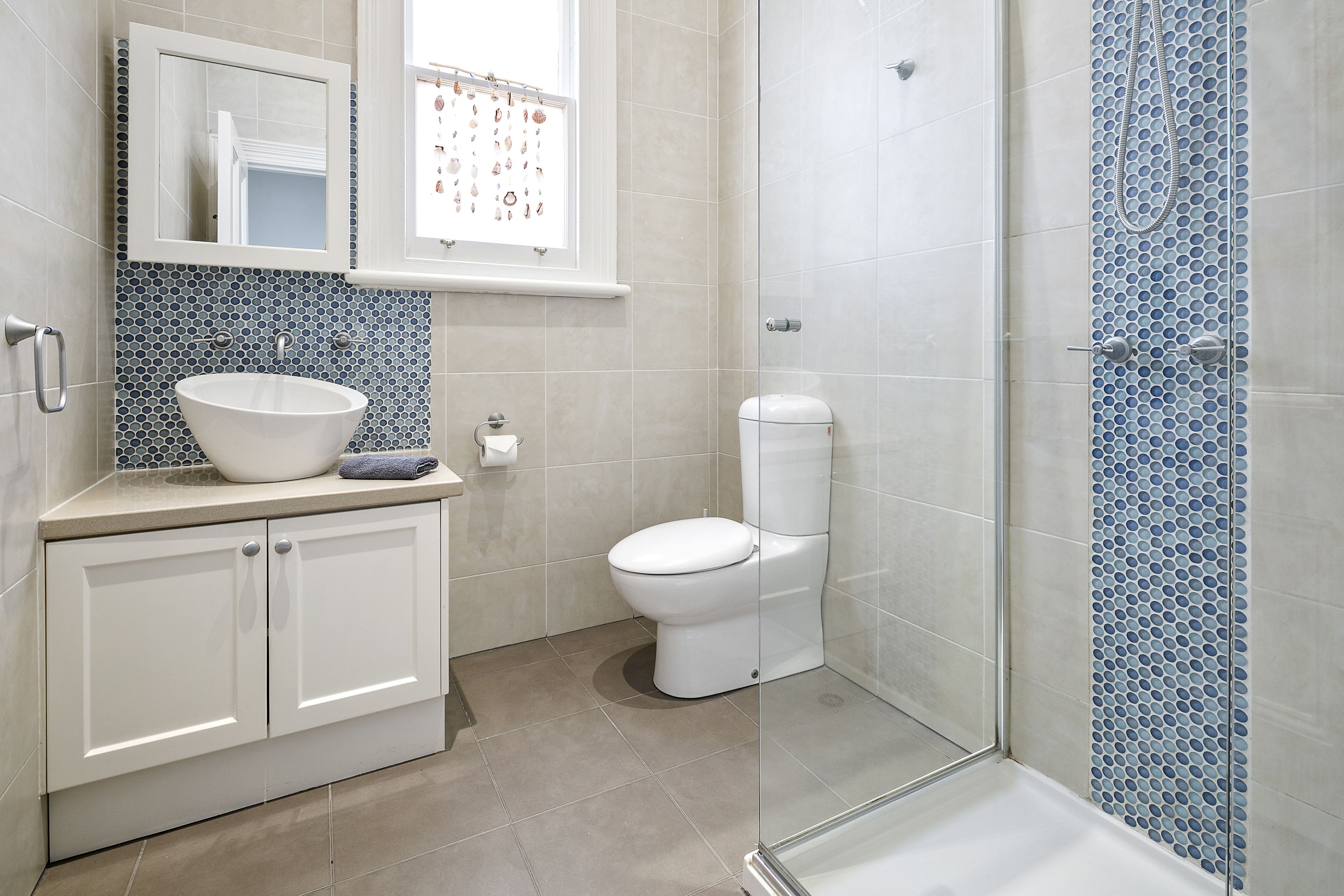 A bathroom with a white sink, toilet, and shower. There is a window above the sink with a decorative mirror.