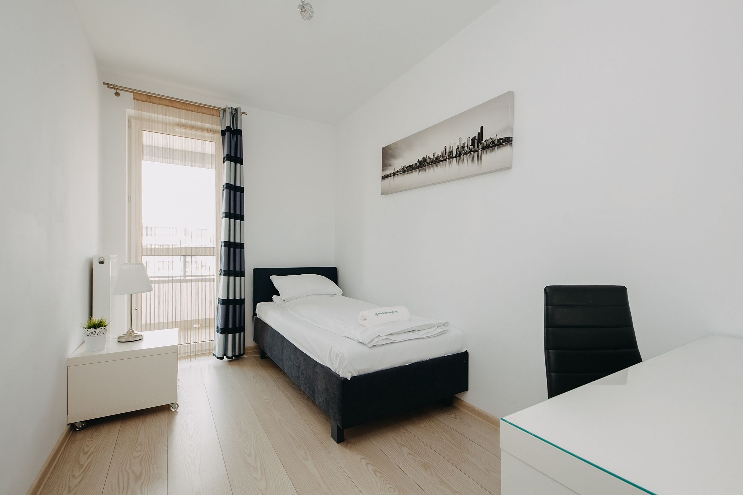 A modern bedroom with a bed, bedside table, lamp, and a cityscape painting on the wall.