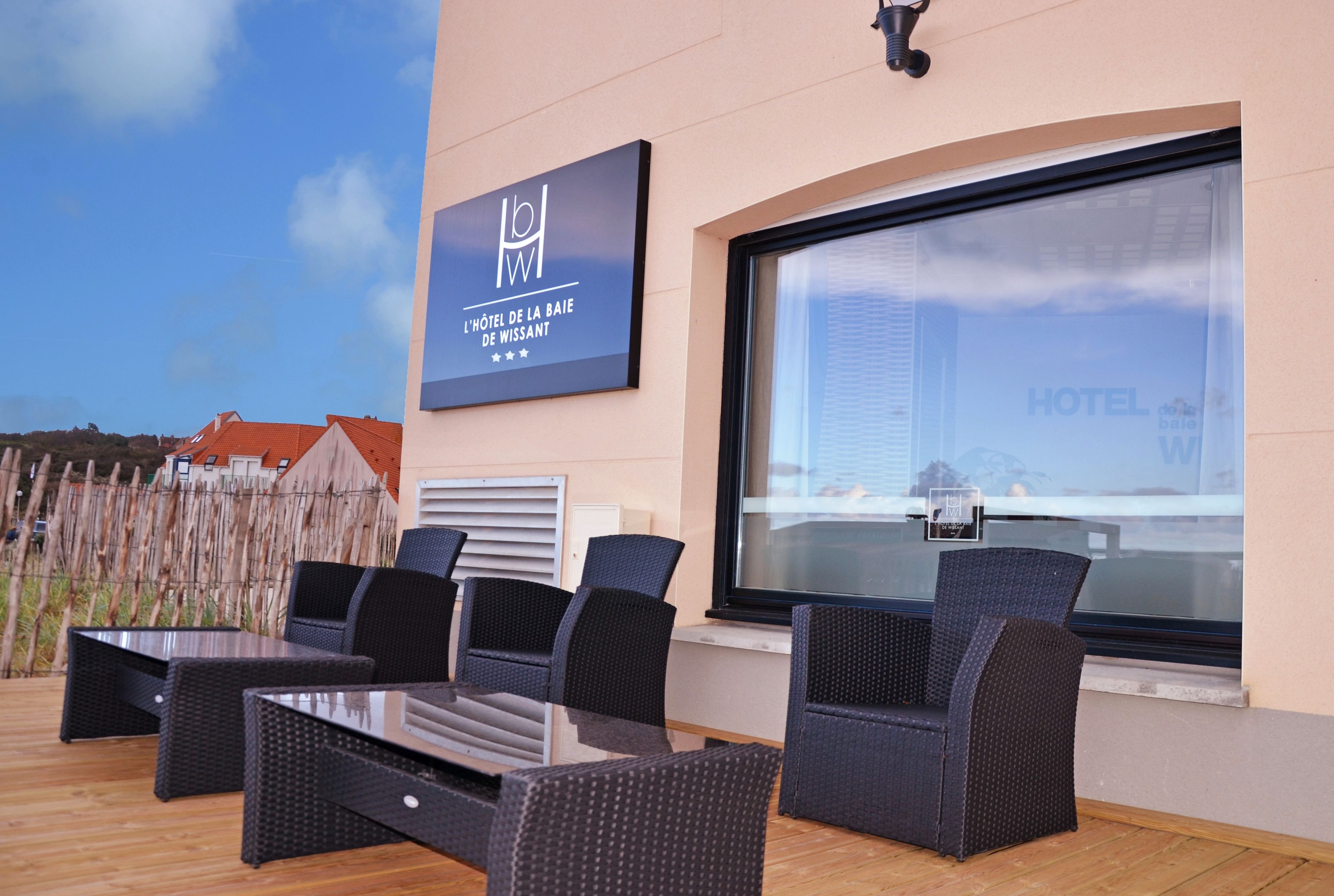 An outdoor seating area with wicker furniture, a glass table, and a sign for 'L'Hôtel de la Baie de Wissant'.