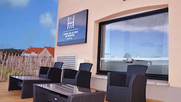 An outdoor seating area with wicker furniture, a glass table, and a sign for 'L'Hôtel de la Baie de Wissant'.