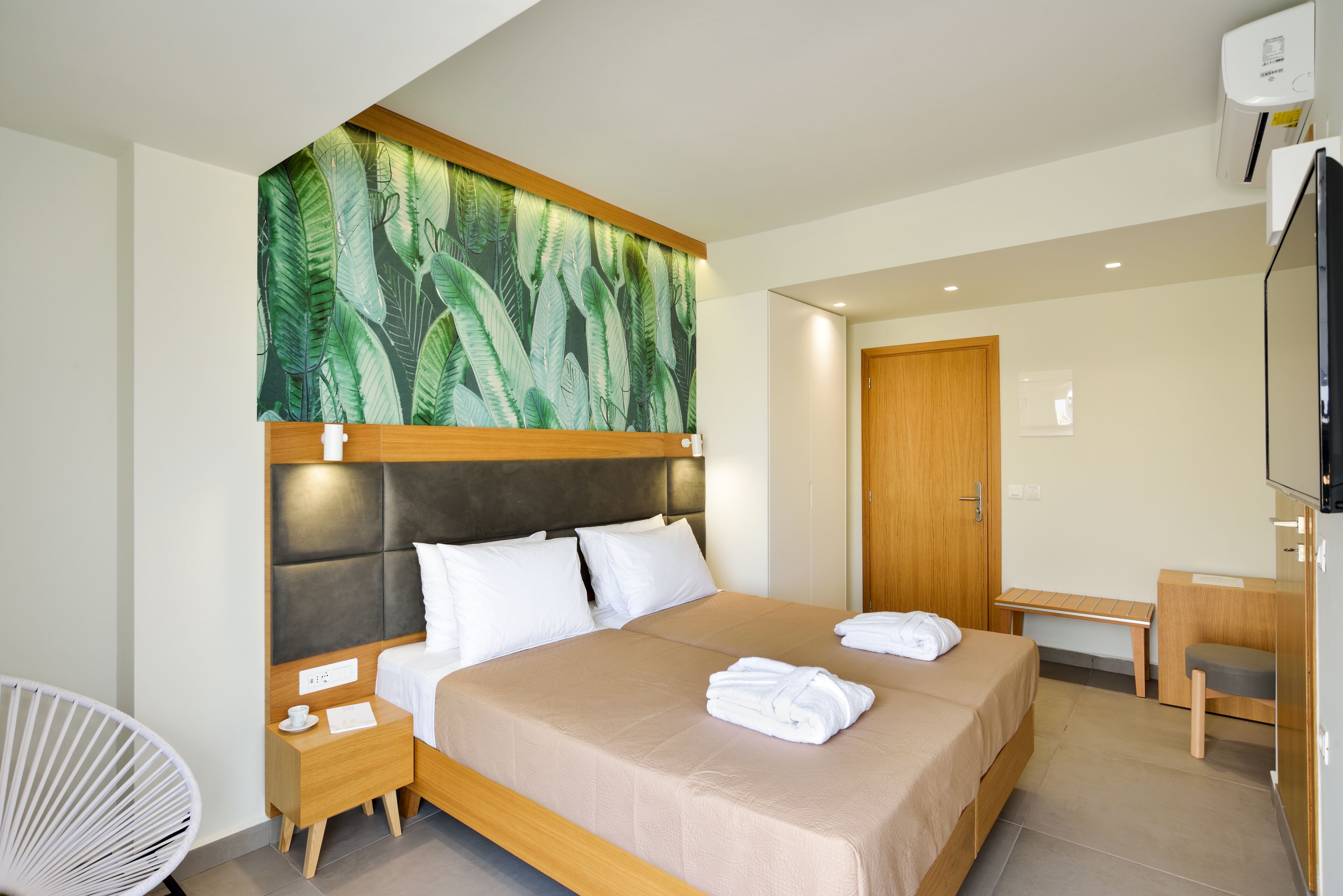 A modern hotel room with a large bed, a wooden headboard, a wall-mounted TV, a wooden desk, and a bench.