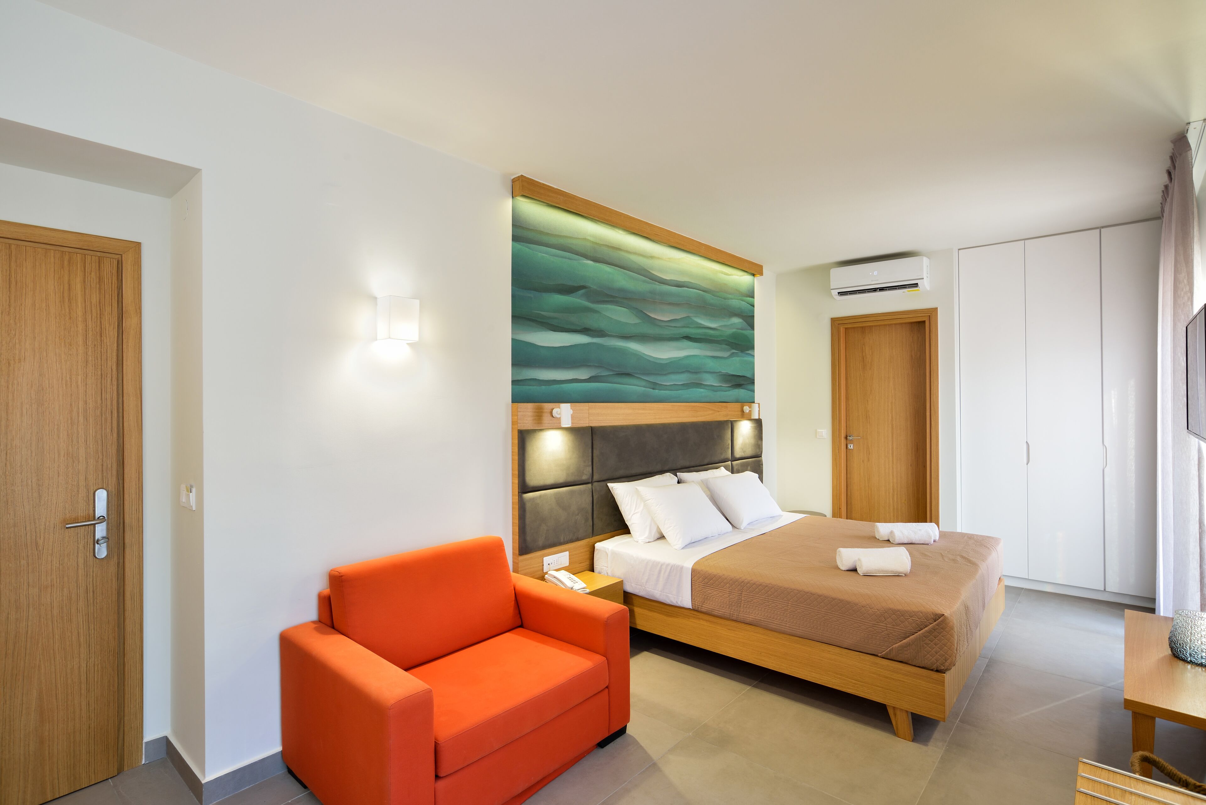 A hotel room with a bed, a wooden headboard, a wall-mounted air conditioner, a wooden door, and a bright orange chair.