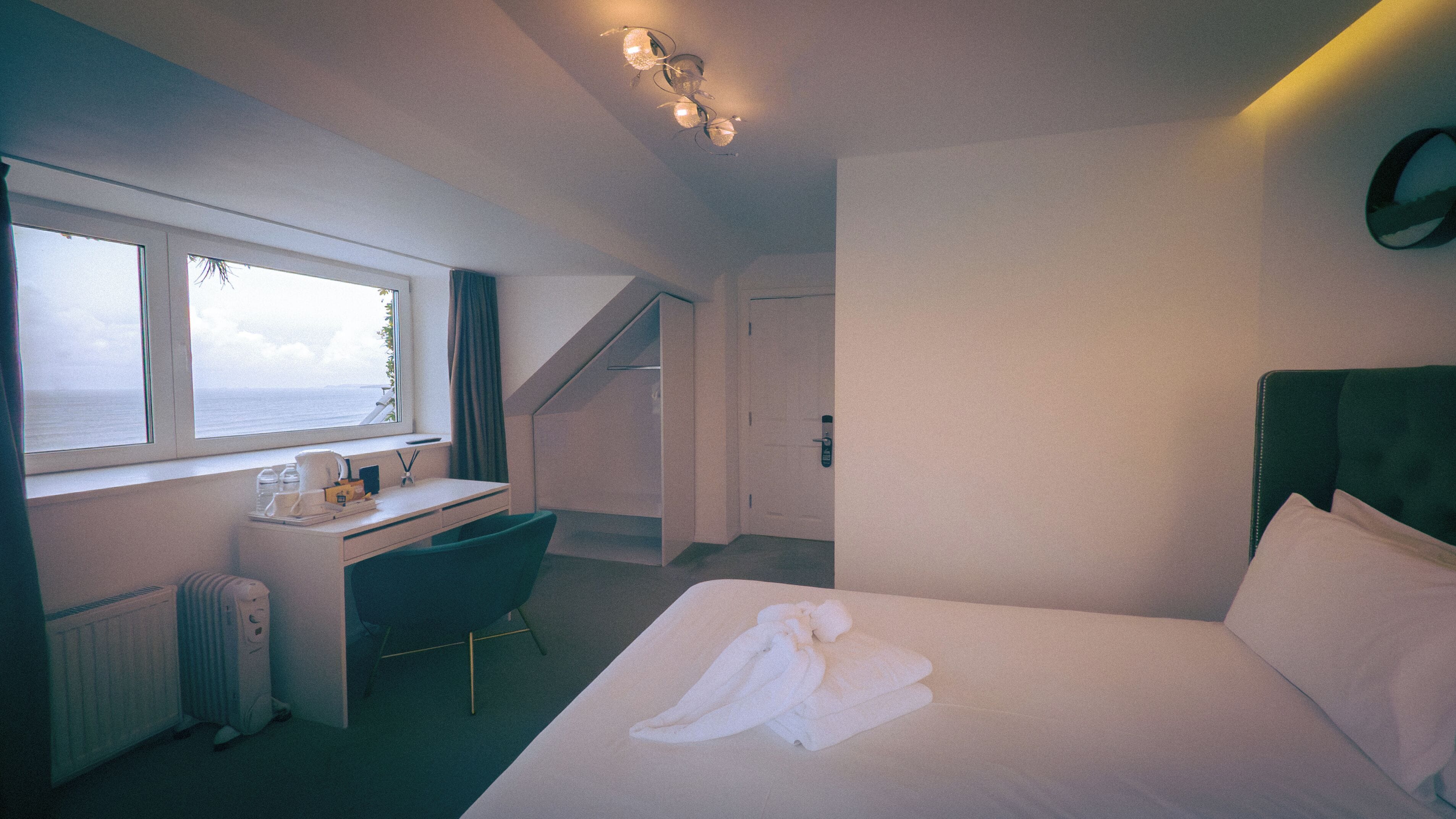 Een hotelkamer met een bed, een bureau, een stoel, een radiator, een raam met uitzicht op zee en een trap.