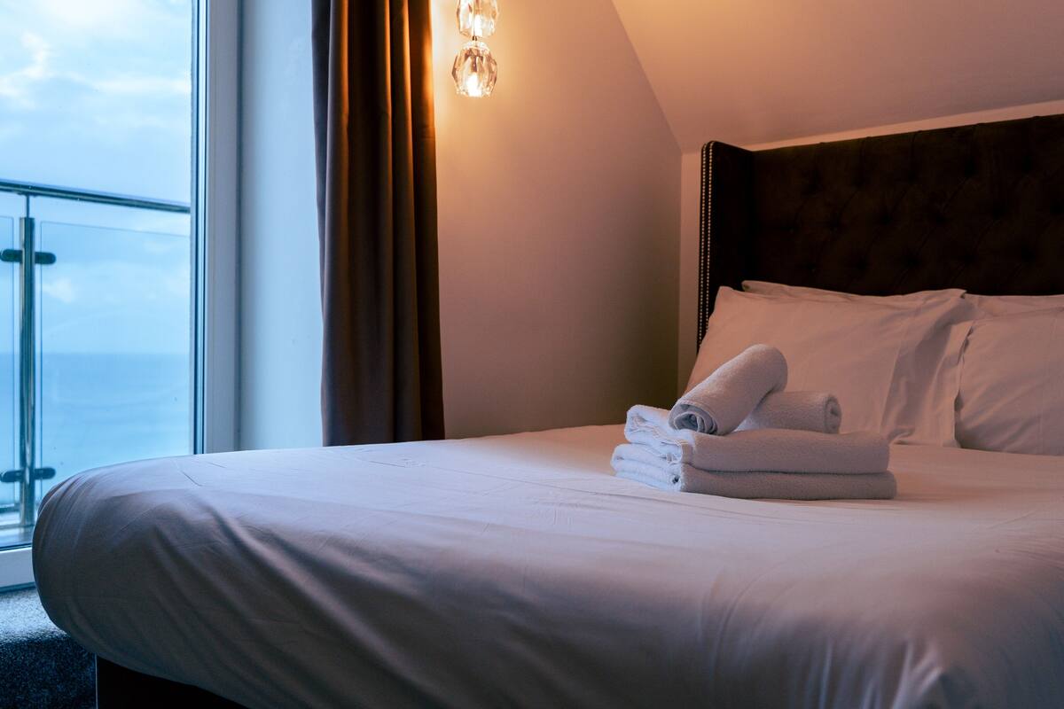 Ein ordentlich gemachtes Bett mit weißen Leinen und zusammengerollten Handtüchern, ein dunkles Kopfstück und ein Fenster mit Vorhängen.