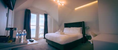 A hotel room with a bed, a desk with a computer, a chair, a window with curtains, and a view of the sea.