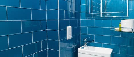 Un baño con azulejos azules, un inodoro blanco, un lavabo, un espejo y un dispensador de papel higiénico.
